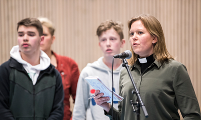Kvinne snakker under et folkemøte. Ungdommer står i bakgrunnen.