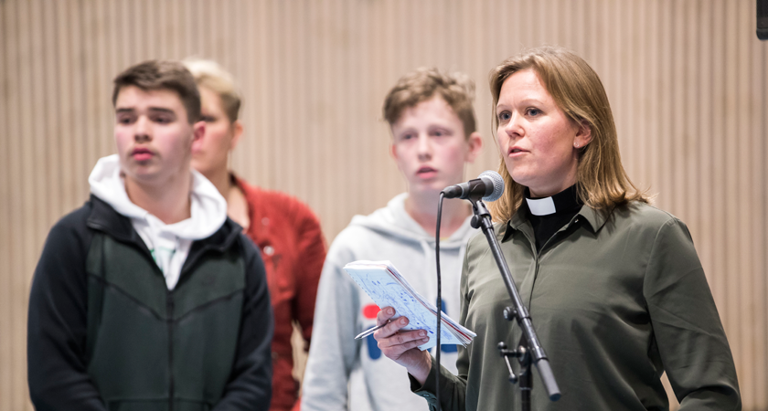 En dame holder et innlegg under et folkemøte. Ungdommer står i bakgrunnen.